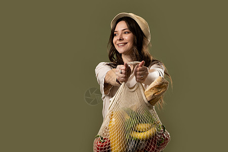 手插口袋的女孩一个身着米色西装 头戴帽子的黑发女孩手里拿着装有水果 蔬菜 果汁和面包的生态袋绳袋 生态友好的概念 时装摄影 零废物概念背景