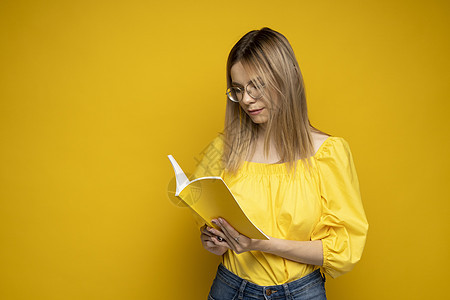 美丽聪明的年轻女孩拿着和阅读孤立在黄色背景上的书 穿着黄色衬衫 戴着眼镜看书的漂亮女人的画像 教育 学习 知识女性大学笔记本女士图片