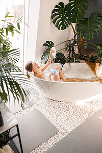 快乐的年轻女子在Loofah海绵上浇浴胶时玩得开心 在现代浴室洗澡 用热带植物装饰淋浴女性浴缸温泉化妆品身体肥皂皮肤呵护风格图片
