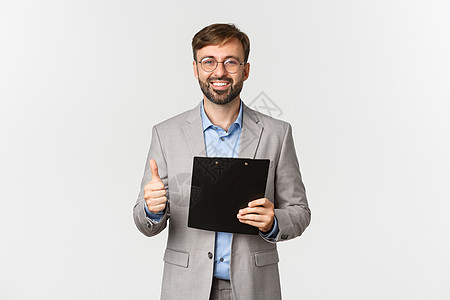 穿着灰色西装和眼镜的满意商务人士肖像 持有剪贴板 在批准时显示拇指抬头 核准好的东西 用白底墨镜进行解码经理办公室管理人员商务喜图片