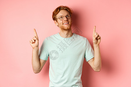梦幻的年轻男人 红头发和胡子 指着手指 看着欢笑起来 仰慕什么 站在粉红色背景上红色学生成人促销眼镜胡须男性购物工作室粉色图片