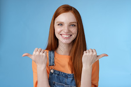 室内拍摄富有魅力 自信 快乐 微笑的红发女人橙色衬衫牛仔工作服指向侧面拇指左右显示选择机会给机会选择 蓝色背景广告护理成人变体手图片