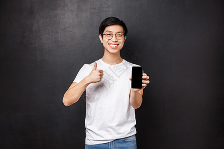 技术 小工具和人的概念 身穿 T 恤和眼镜的自信可爱的亚洲男人 推荐网上商店或手机应用 竖起大拇指和智能手机显示屏图片