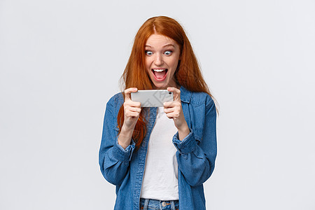 兴奋 快乐和惊讶的漂亮红发女学生击败分数 赢得比赛 挑战智能手机游戏 水平拿着手机 观看派对上的搞笑视频 看起来很惊讶图片