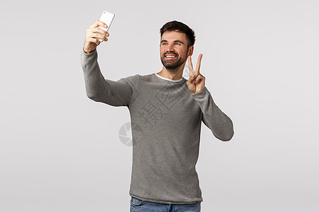乐观 无忧无虑 快乐 有魅力的男性 留着灰色毛衣的胡子 举着智能手机自拍 做出和平标志 微笑着 在网上给朋友视频通话 白色背景图片