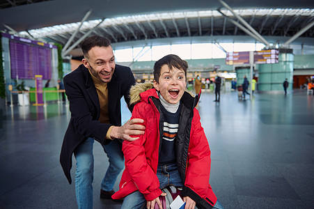 开朗快乐的父亲在国际机场的出发大厅里骑着他的儿子在行李上 爸爸和儿子在等待护照 海关检查和登机时一起玩图片