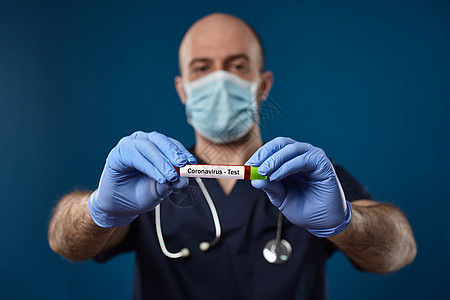 戴着面具 蓝色手套 医疗服的医生 拿着有绿色盖帽的红色实验室试管在手上 蓝色背景 冠状病毒COVID19 特写男人成套肺炎测试生图片