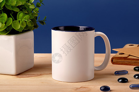 木制桌面上的白色陶瓷杯子 旁边是散落的玻璃石 锅中的绿色植物和棕色笔纸 蓝色背景图片