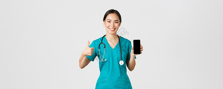 Covid19 医护人员和在线医学概念 亚洲年轻女医生的画像 身着磨砂膏的医生竖起大拇指和智能手机屏幕 推广应用 白色背景看门人图片