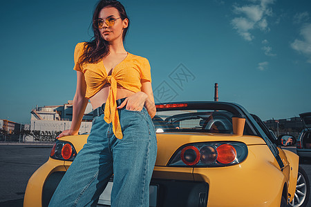 穿着蓝色牛仔裤 橙色上衣和太阳镜的女孩正靠在黄色敞篷车上 在停车场的后备箱上放着纸杯饮料 复制空间图片