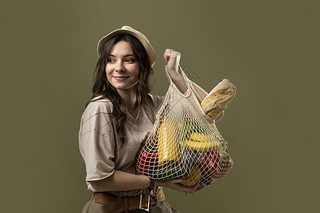 穿着浅色夏装的年轻女子微笑着 带着一个装满蔬菜的网眼生态袋 绿色工作室背景上的绿色蔬菜 可持续的生活方式 生态友好的概念 零浪费图片