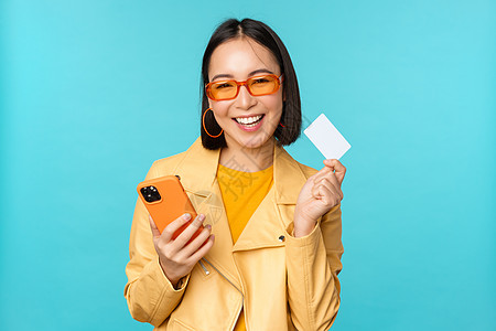 网上购物 戴墨镜 出示信用卡和使用智能手机 在网上支付 购物 站在蓝色背景上的时尚亚洲年轻女性企业家工作黑发技术太阳镜促销电话办图片