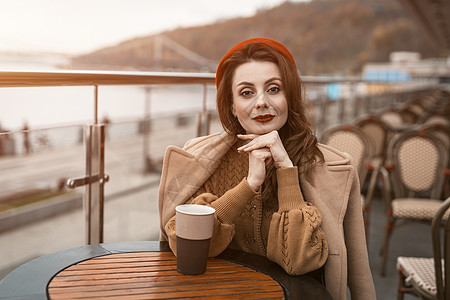 可爱的法国年轻女子坐在餐厅露台上 拿着咖啡杯看着相机 户外穿着秋衣和红色贝雷帽的时尚年轻女性肖像桌子咖啡店闲暇食物女人女孩商业城图片