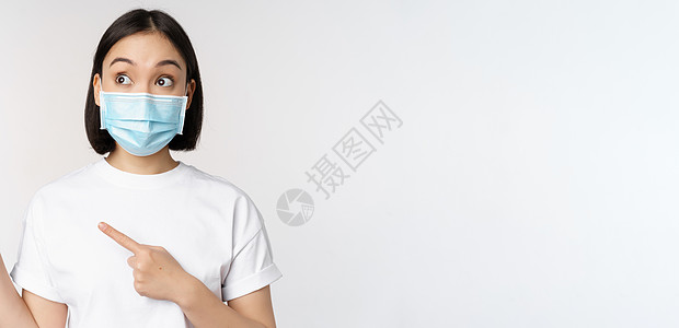带着医疗面罩的韩国年轻女医务人员用左手指向左边 看标志 张贴广告或标语 站立在白色背景上疫苗保健面具女性学生商业购物技术企业家情图片