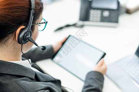 耳机包装一名佩戴头盔的商业妇女坐在办公桌旁时持有数字平板电脑创业公司屏幕药片职场互联网机动性商务企业家套装背景