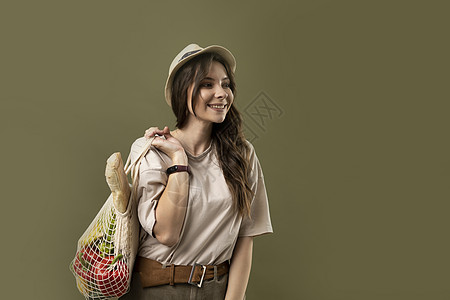 手插口袋的女孩穿着浅色夏装 带着装满蔬菜的网眼生态袋 在绿色工作室背景下的相机里看着绿色植物的年轻女子微笑着 可持续的生活方式 生态友好的概念背景