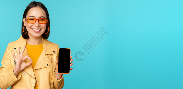 微笑的韩国女性展示手机应用程序界面 智能手机应用程序 手机推荐 站在蓝色背景上办公室情绪工作太阳镜工作室情感商业企业家成人购物图片