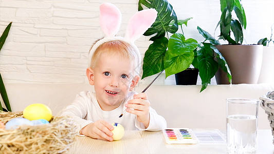 一个小兔子耳朵里的小男孩 热情地用油漆给复活节鸡蛋涂彩画 宗教假日概念 你自己做 家庭休闲 东边猎鸡假期幼儿园戏服教育篮子画笔孩图片