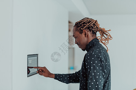 微笑的非洲裔美国人使用现代智能家居系统 墙上的控制器 积极的年轻人在恒温器上切换温度或激活公寓的安全警报创新互联网技术笔记本家庭背景图片