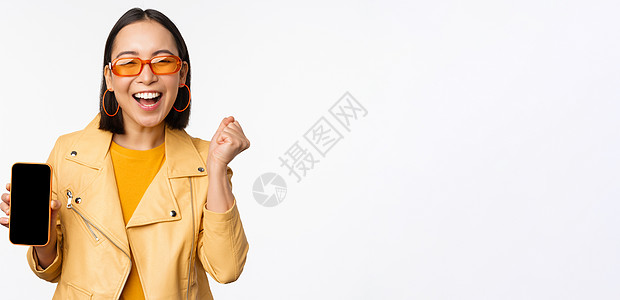 快乐的亚洲女孩在太阳镜中 展示移动电话屏幕 智能手机界面 笑笑和微笑 庆祝 站在白色背景上站立促销工作学生电话广告黑发情感商业购图片