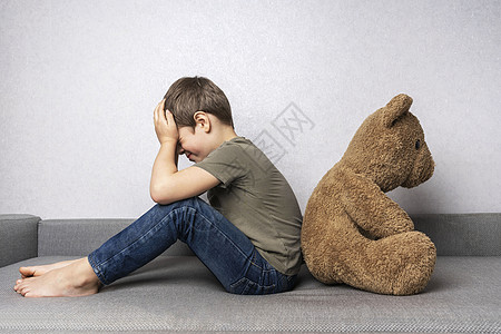 一个沮丧的学龄前男孩坐在沙发上 背靠着一个大泰迪熊哭泣摄影房间挫折压力幼儿园男生朋友情感愤怒图片