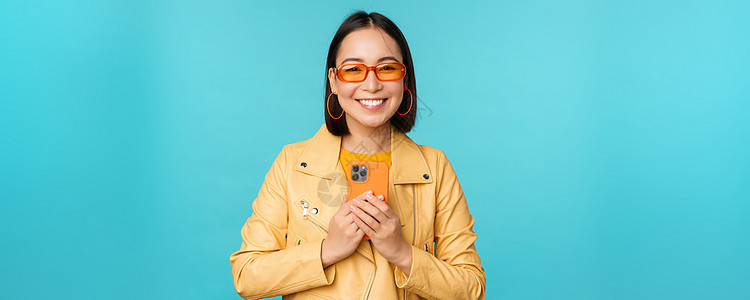 戴着墨镜 手持智能手机 用手机拍照 微笑 录音 站在蓝色背景上的时尚 美丽的中国女性形象电话商业成人企业家黑发促销学生情绪办公室图片