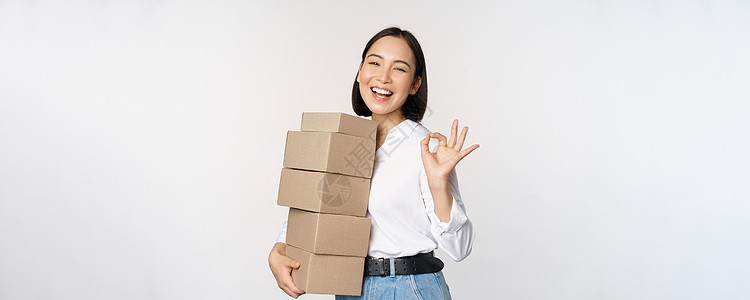 美丽幸福的女人的肖像携带订单的盒子 带运货并显示好的标志 白色背景的女商务人士商业成人工作室手臂商务冒充商品员工产品售货员图片