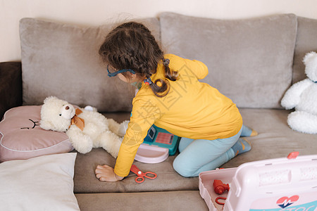 快乐的小女孩在她的房间里扮演医生 可爱的三岁女孩花时间玩耍 戴眼镜的女孩黄色上衣和蓝色紧身裤 乌克兰可爱的孩子 她的孩子厨房的背图片