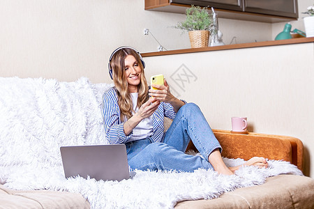 远程学习在线教育和工作 戴着耳机进行视频通话的女人 快乐和微笑的女孩在沙发上在家庭办公室工作 喝着咖啡 使用电脑和手机图片