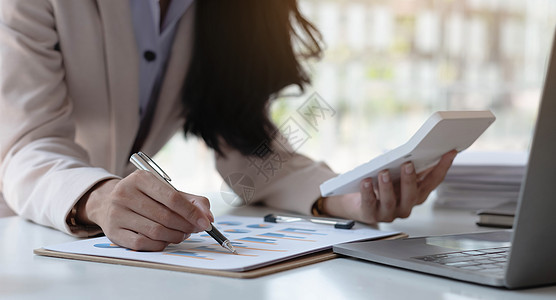 一名女商务人士手握笔 用办公室装有计算器眼镜笔记本电脑的图表指着一张图图片
