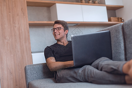 年轻人用笔记本电脑 躺在客厅沙发上写作成人房间男性长椅蓝图公寓技术网络闲暇图片