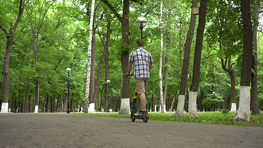 一名穿衬衫和短裤的欧洲青年骑着电动摩托车在公园里行驶骑术成人轮子游乐电话树木电气车辆驾驶技术图片