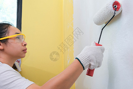 戴护目镜的年轻女装饰师用滚筒涂墙壁 修理和室内装饰背景图片