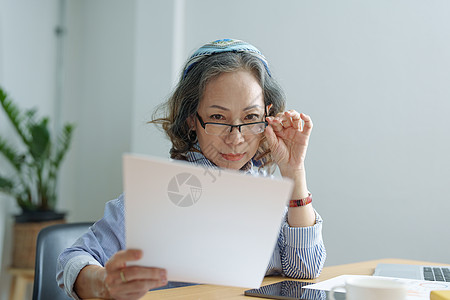 老年妇女手持重要文件核实纸上笔记真伪的画像桌子女士银行女性金融账单审计预算退休代理人图片