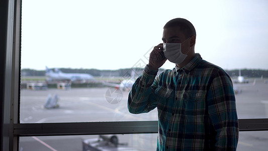 一名戴医疗面具的年轻人在机场窗户的背景下通过电话交谈 背景是空中飞机(见A/C 5/49/70 附件)图片