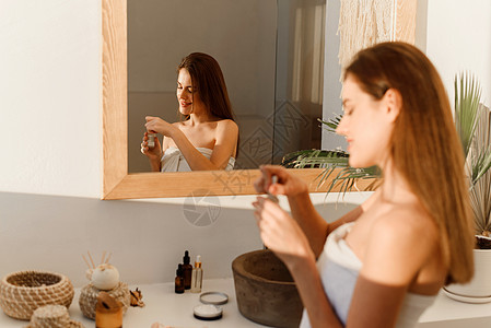 面部皮肤护理 使用透明质酸面部精华液 保湿和护肤品的迷人女性站在现代室内浴室的镜子附近 日常美容程序产品吸管皮肤科液体化妆品卫生图片