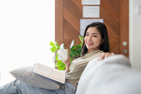 在沙发上笑着的亚洲女人 看着一本看相机的书 早上喝咖啡图片