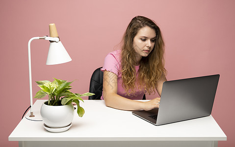 身穿粉红色 T 恤的年轻自由职业者卷发女性使用笔记本电脑工作 在一个项目上工作 自由职业者工具衣服桌子网络互联网人士撰稿人椅子工图片