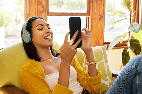 西班牙裔女性在明亮的客厅里舒适放松地使用智能手机 戴着耳机听音乐 一位坐在椅子上的年轻女性在家里用手机发短信图片