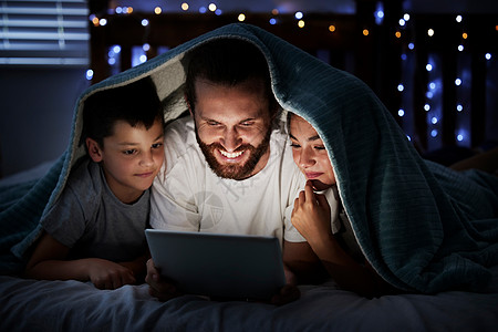 快乐的白人家庭单身父亲有两个孩子 晚上在黑暗中使用数字平板电脑躺在毯子下 他们的脸被设备屏幕灯照亮 父亲在晚上与女儿和儿子一起阅图片