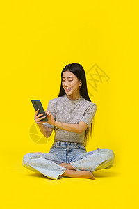 界面展示手持电话的亚洲女孩在地板上用手机发短信或买网上玩耍游戏 孤立于黄色背景 移动电话应用程序广告产品布置 复制空间 (续)背景
