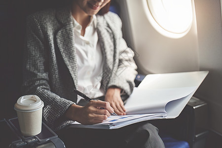 一位成功的亚洲女商务人士或女企业家在飞机上穿着正式西装 坐在商务舱座位上 并在飞行期间使用带有文件的笔进行工作电脑管理人员裁剪航图片
