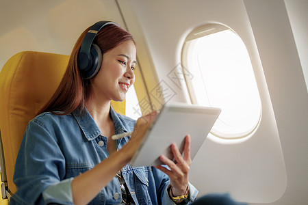 飞机撒药一位坐在经济舱靠窗座位上的亚洲女性在飞机飞行 旅行概念 假期 放松期间使用平板电脑并听器乐的迷人肖像窗户快乐技术企业家旅游享受药背景