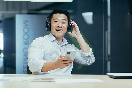 年轻的亚洲商务人士在耳机和电话中听音乐 休息了一下工作 笑声商业人士互联网领导者职业电脑笔记本男人男性享受图片