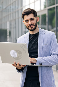 留着胡须的年轻成功商务人士自由职业者正站在现代办公室附近 看着镜头微笑 男人正在他的笔记本电脑上工作图片