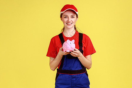 工作妇女站在小猪银行的手边 带着快乐的表情看着相机图片