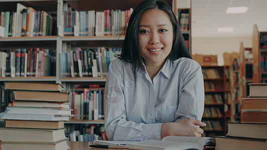 年轻美丽的亚洲女学生坐在桌旁 图书馆里成堆的教科书看着相机的画像 她笑得很积极学习阅读家庭作业大学青少年教育学校书架快乐女士图片