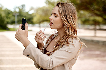 是时候做出这些动作了 但首先要化妆 一个年轻的女商务人士在城市背景下使用智能手机涂口红图片