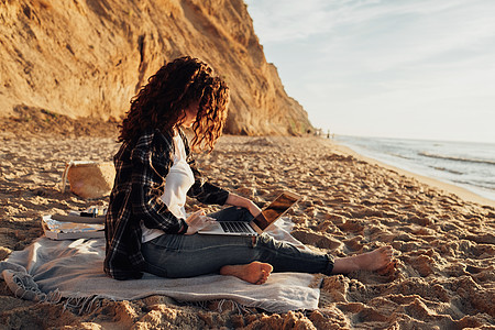 坐在月亮上月亮黎明坐在海边时 身穿笔记本电脑的卷毛黑发妇女 背景上有美丽的海岸线海洋互联网自由职业者卷发女孩格子海岸野餐毯子女性背景