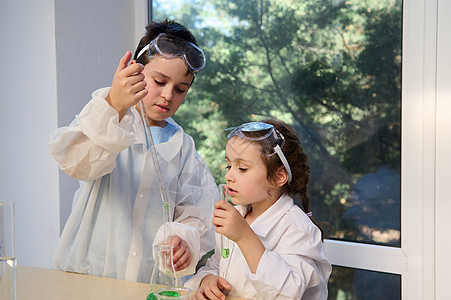 身穿白色实验室大衣和安全护目镜 在化学类中进行科学实验的适龄儿童 男孩和女孩图片